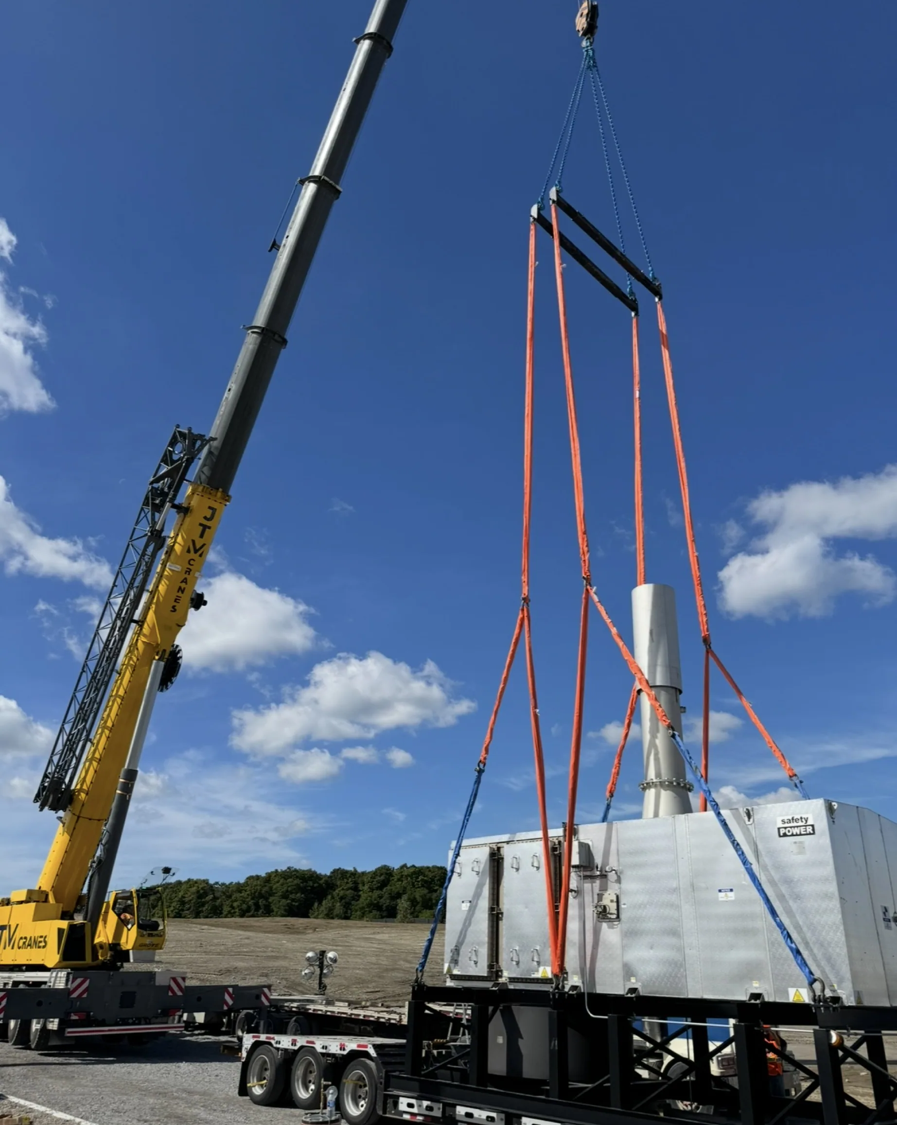 JTM Cranes crane lifting a large machinery unit off a flatbed trailer with spreader rigging
